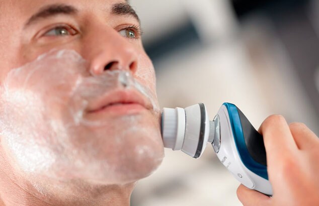 Close-up of a man with shaving foam on his face using a white, blue, and grey electric shaver.