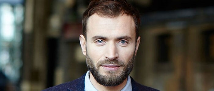 Brown-haired man in suit with a defined jawline, accentuated by a carefully-shaped full beard.