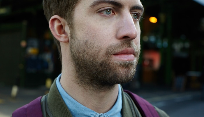 Brown-haired man with 1cm beard covering face and neck standing in a street wearing purple rucksack.